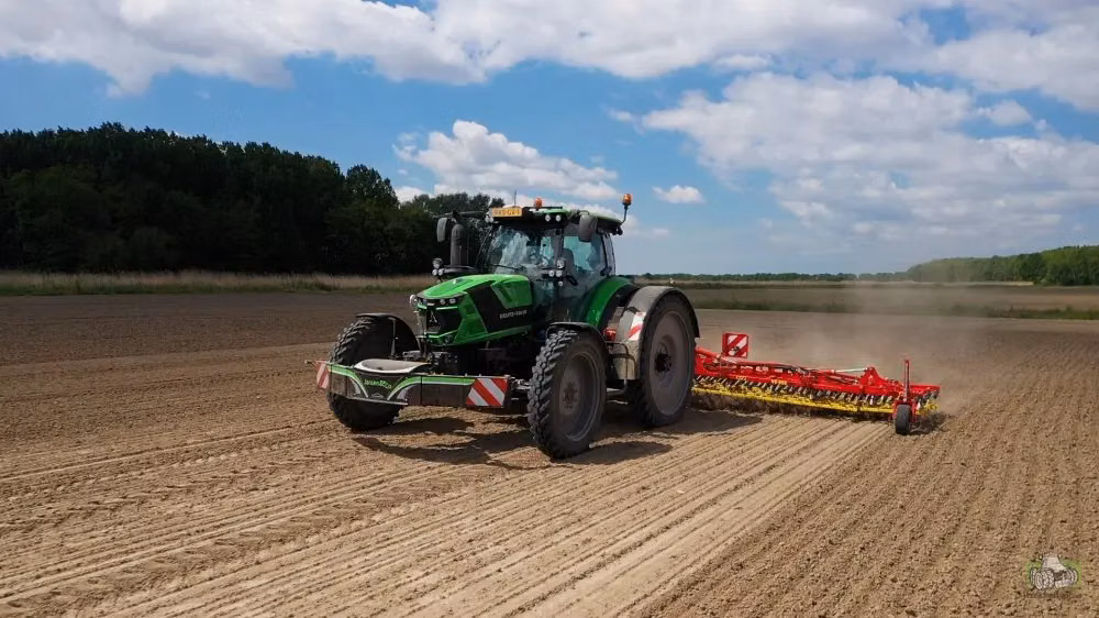 Green tractor with a front weight bumper and a red cultivator attachment working on a plowed field under a blue sky