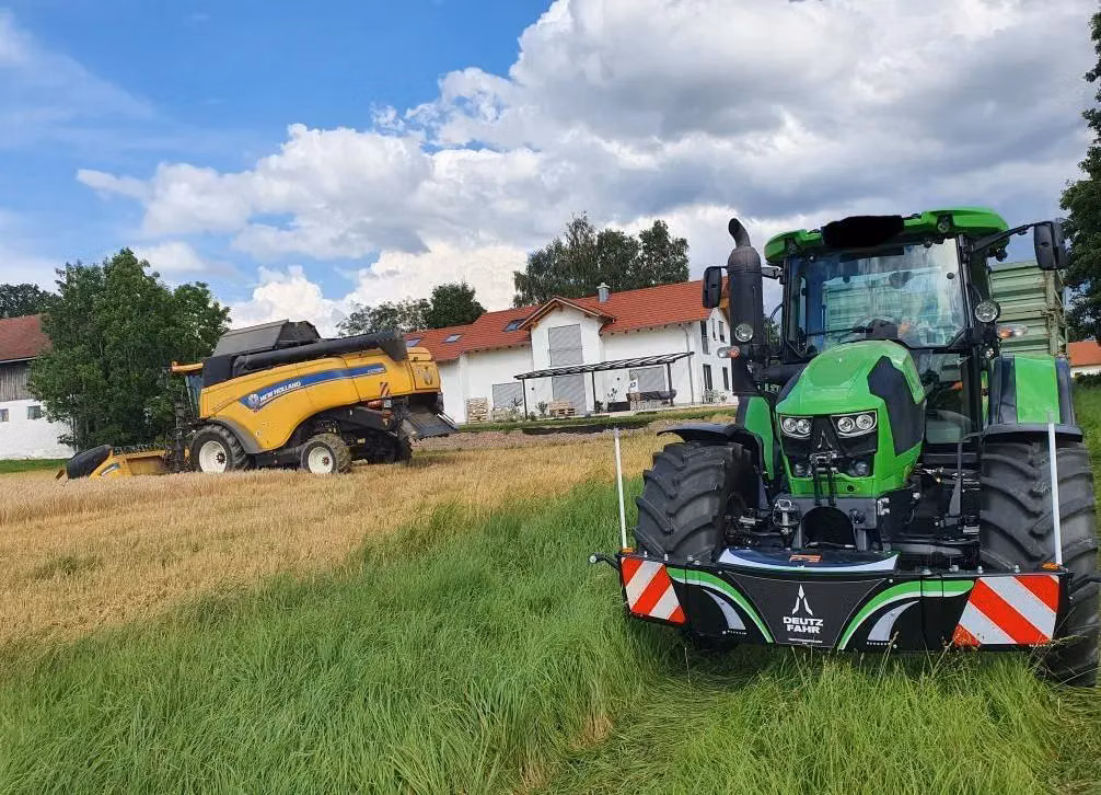 Green tractor with a Deutz Fahr front weight bumper in a grassy field, with a yellow harvester and farm buildings behind