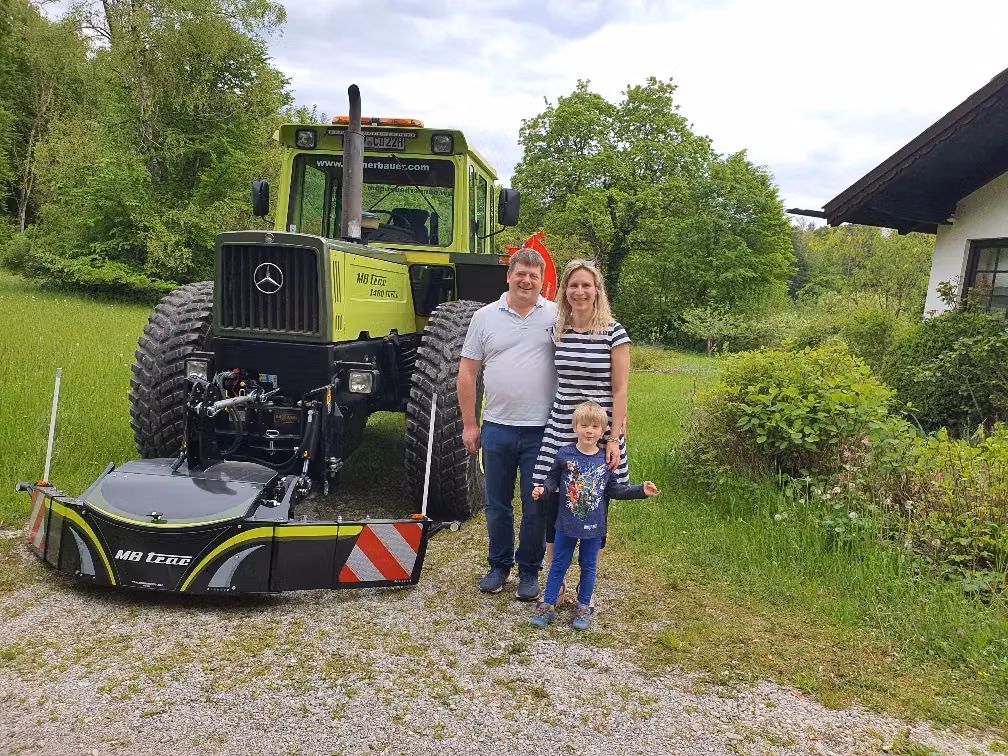 A family stands in front of a green MB trac tractor fitted with a large front weight bumper on a gravel path.