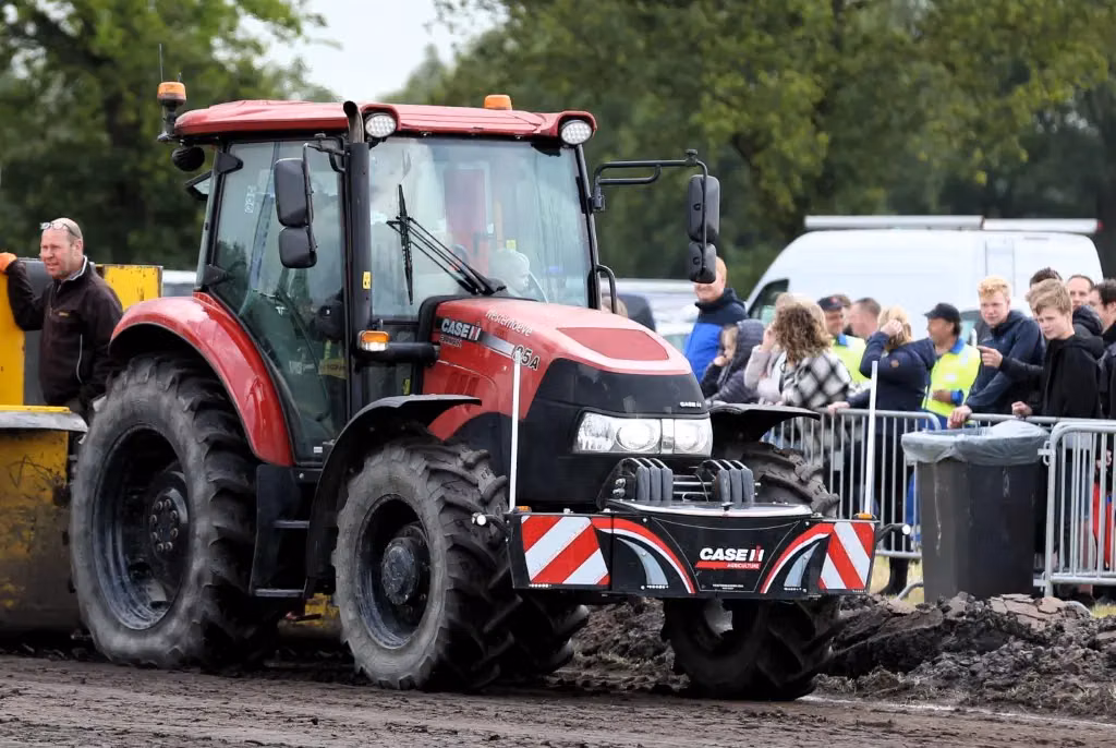 Red tractor with a CASE IH branded front weight bumper, driving on muddy ground near a group of spectators and barriers