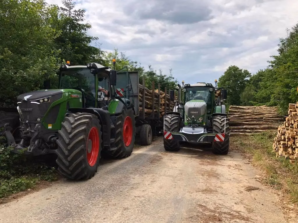 Two green tractors with front weight bumpers are parked on a rural road, each hauling trailers loaded with logs