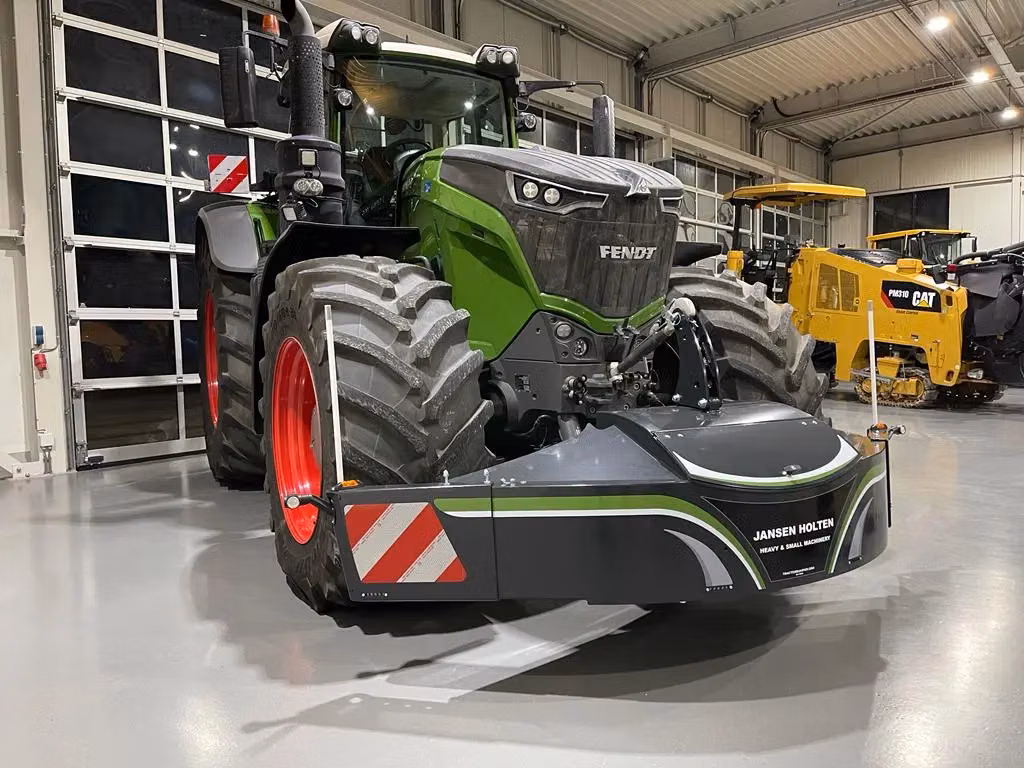 A green tractor with red wheels is fitted with a large black front weight bumper labeled Jansen Holten in an indoor showroom