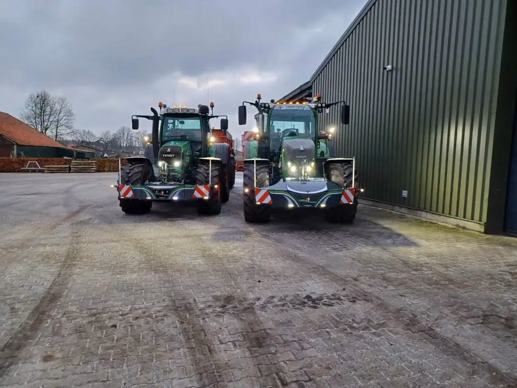 Two tractors with LED front weight bumpers and reflective markings are parked side by side near a large building