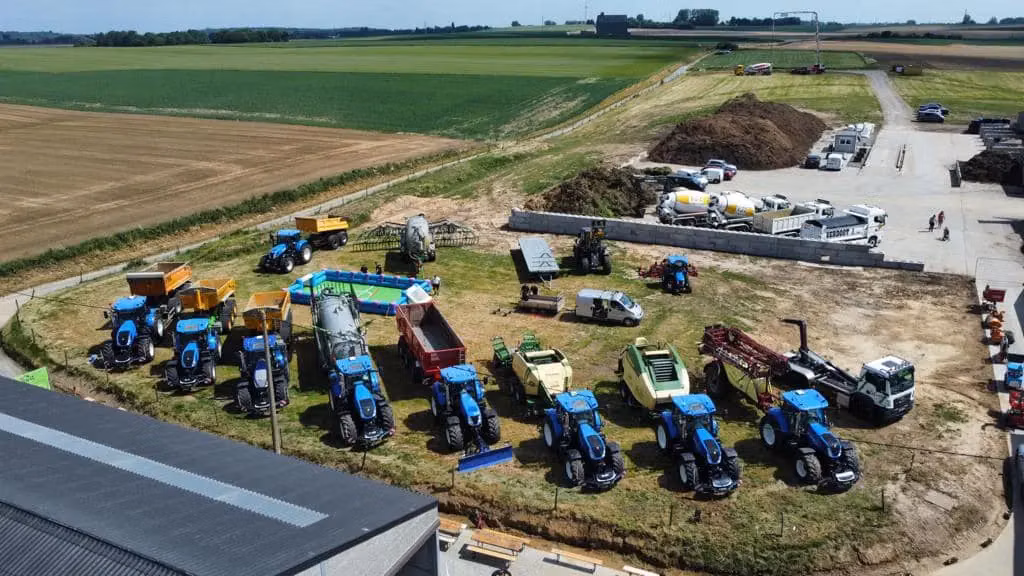 Several blue tractors equipped with front weight bumpers are parked in a row on a farm with various other machinery