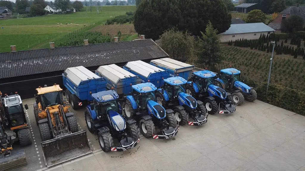 Five blue tractors with front weight bumpers are parked in a row by blue trailers alongside other farm machinery