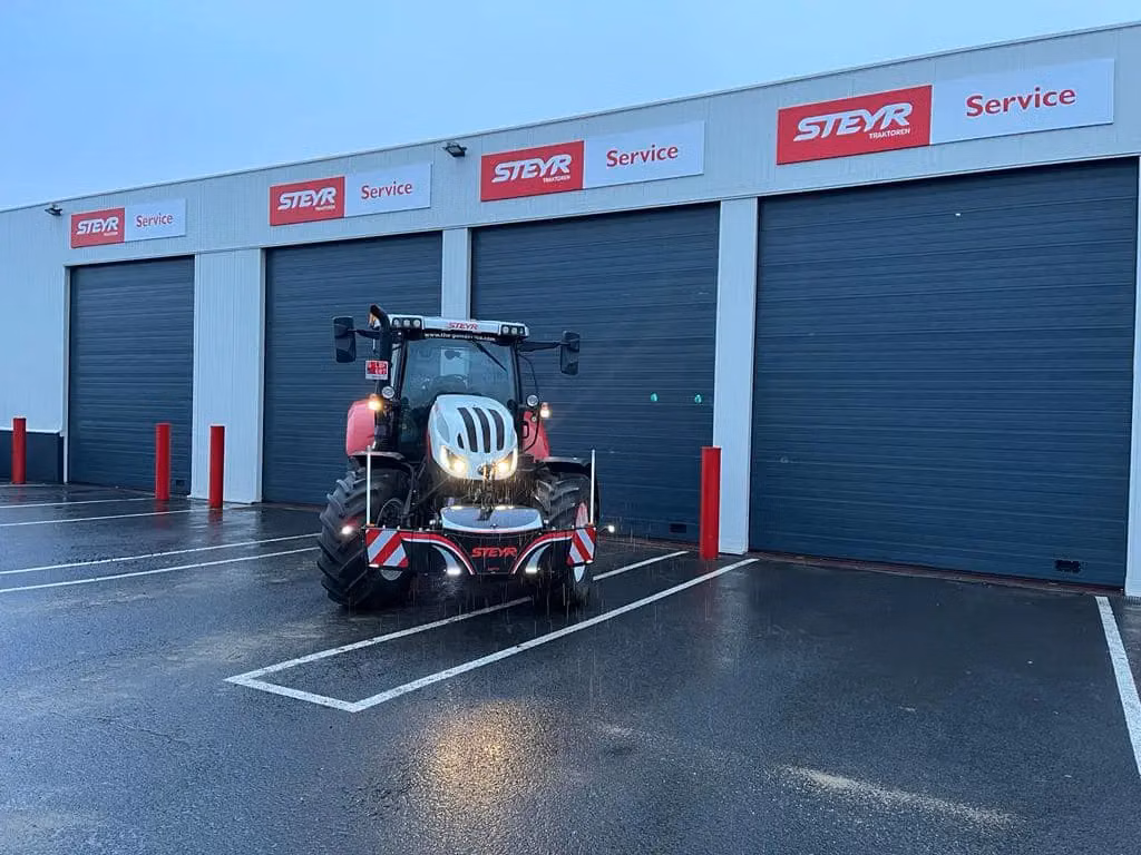 A tractor with a front weight bumper is parked in front of a building with multiple service garage doors.