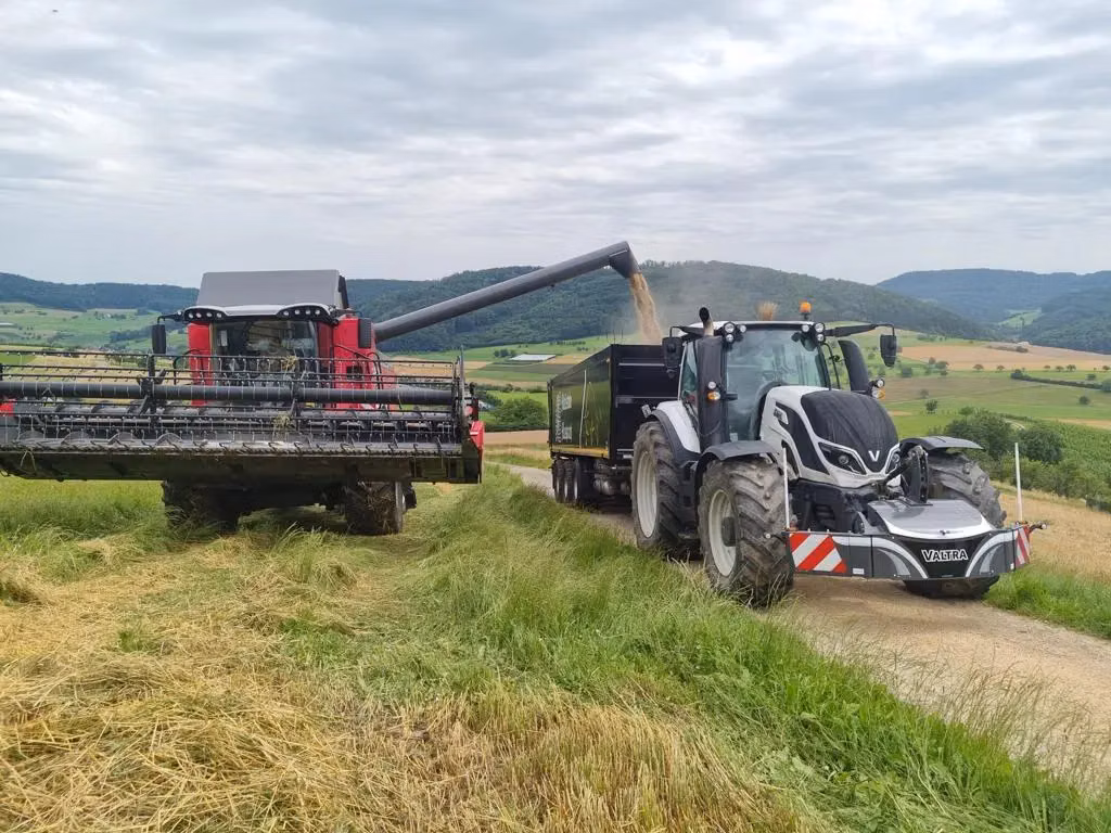A tractor with a front weight bumper labeled Valtra is parked beside a combine harvester unloading grain into a trailer
