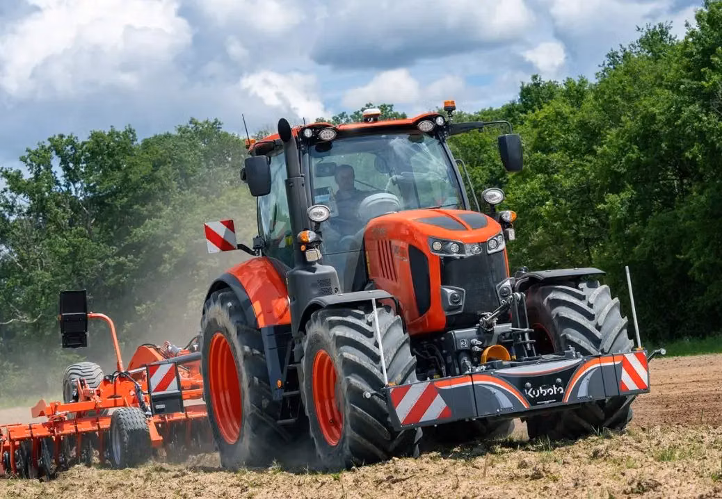 A large orange tractor with a front weight bumper and reflective stripes working in a field with attached equipment