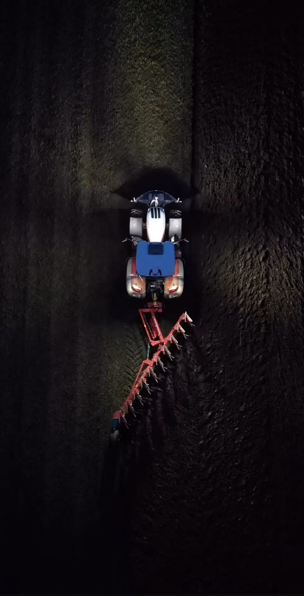 Tractor with a front weight bumper and mounted plough working on a field at night, viewed from above