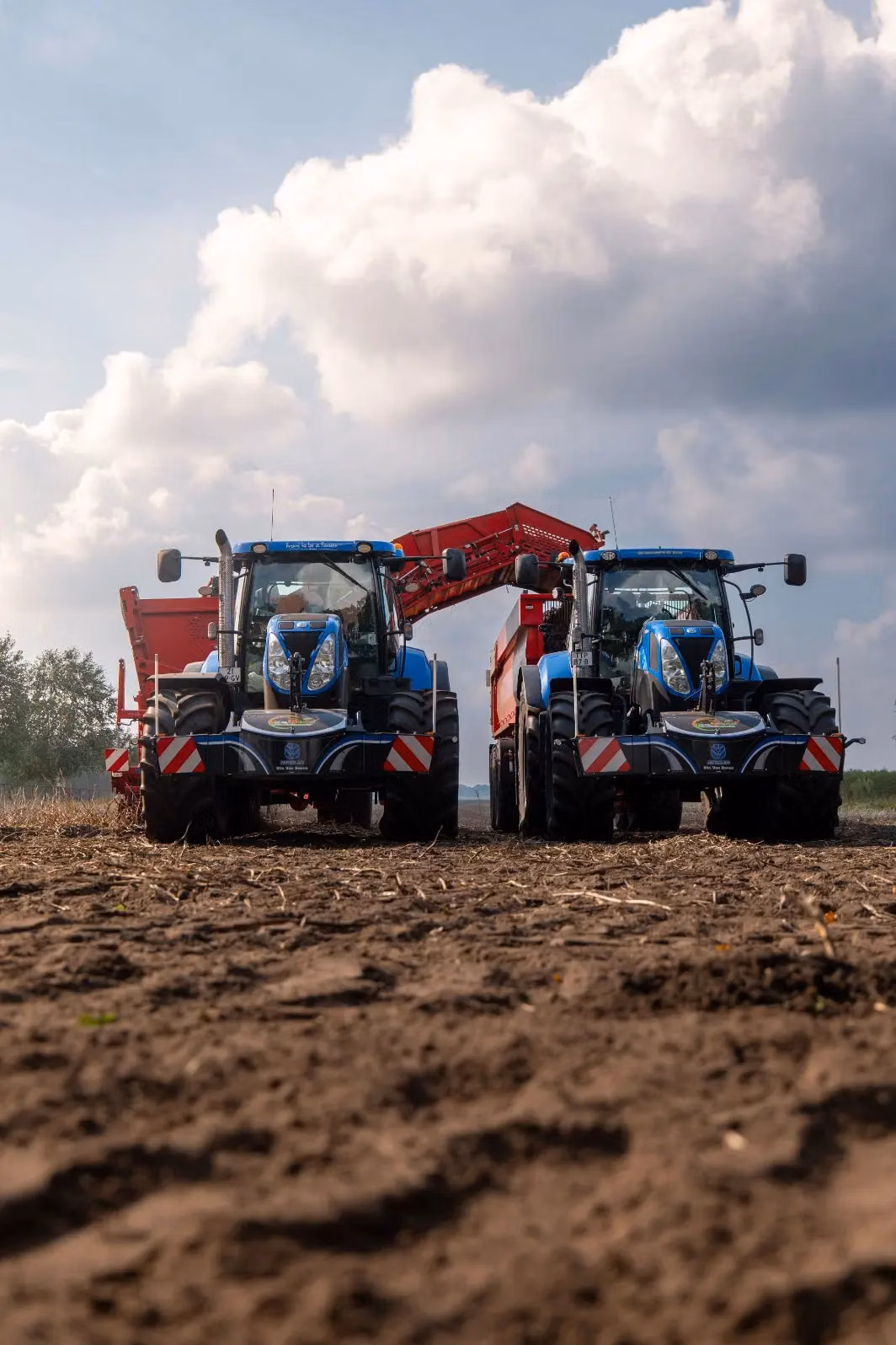 Two blue tractors with front weight bumpers are parked side by side on a dirt field under a cloudy sky