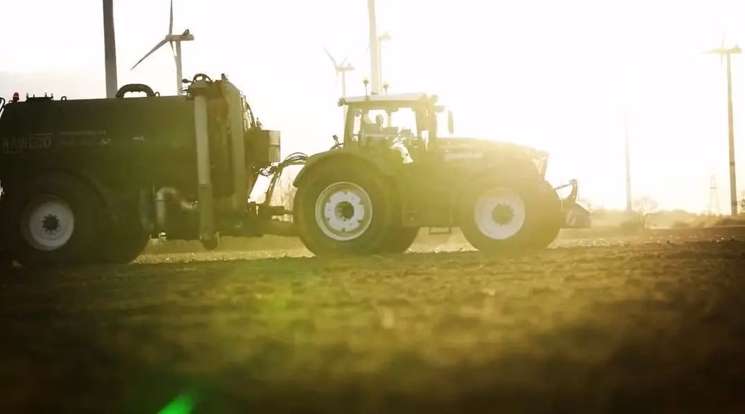 A tractor with large white wheels pulls a tank trailer across a field at sunset with wind turbines in the background