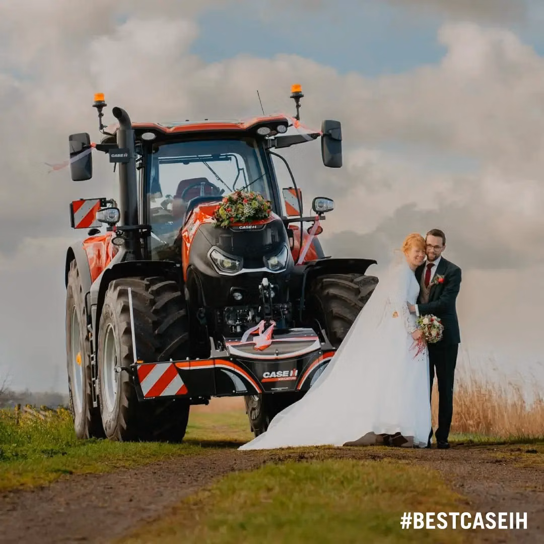 A decorated red tractor with a front weight bumper is parked on a dirt path next to a bride and groom