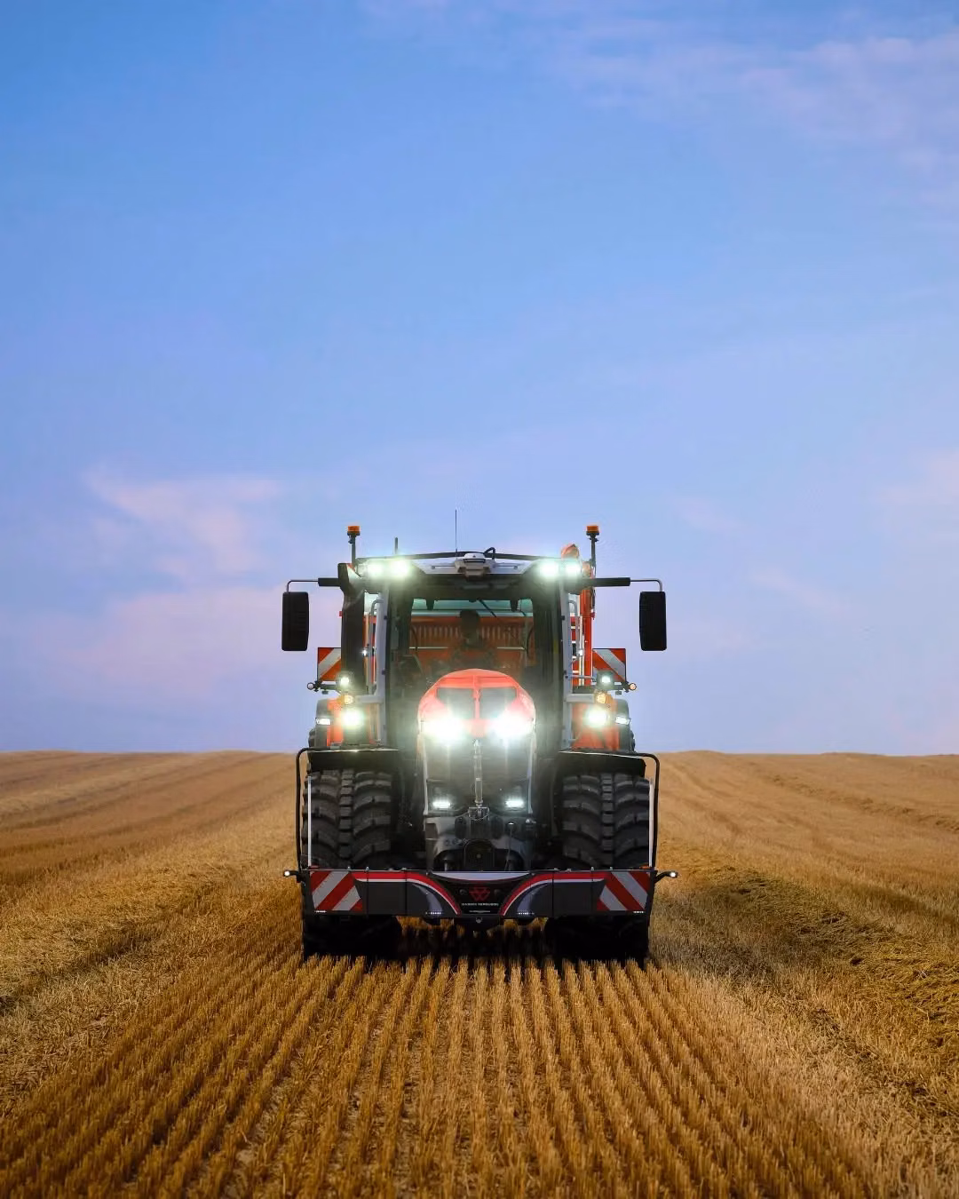 Tractor with front weight bumper and headlights on, driving through a harvested field under a blue sky
