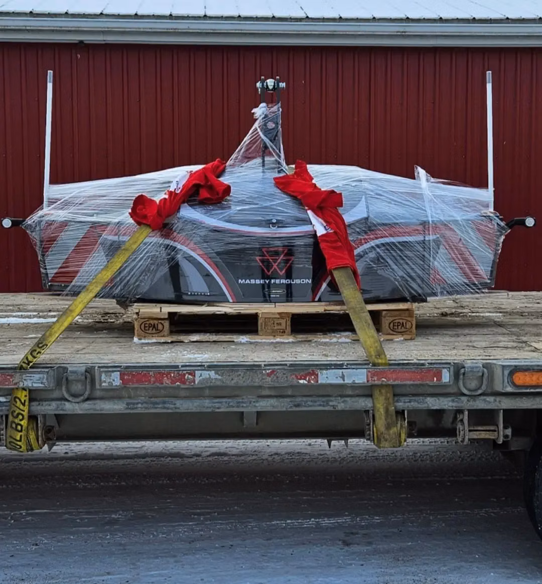 A Massey Ferguson front weight bumper wrapped in plastic with red bows is secured on a pallet and flatbed trailer