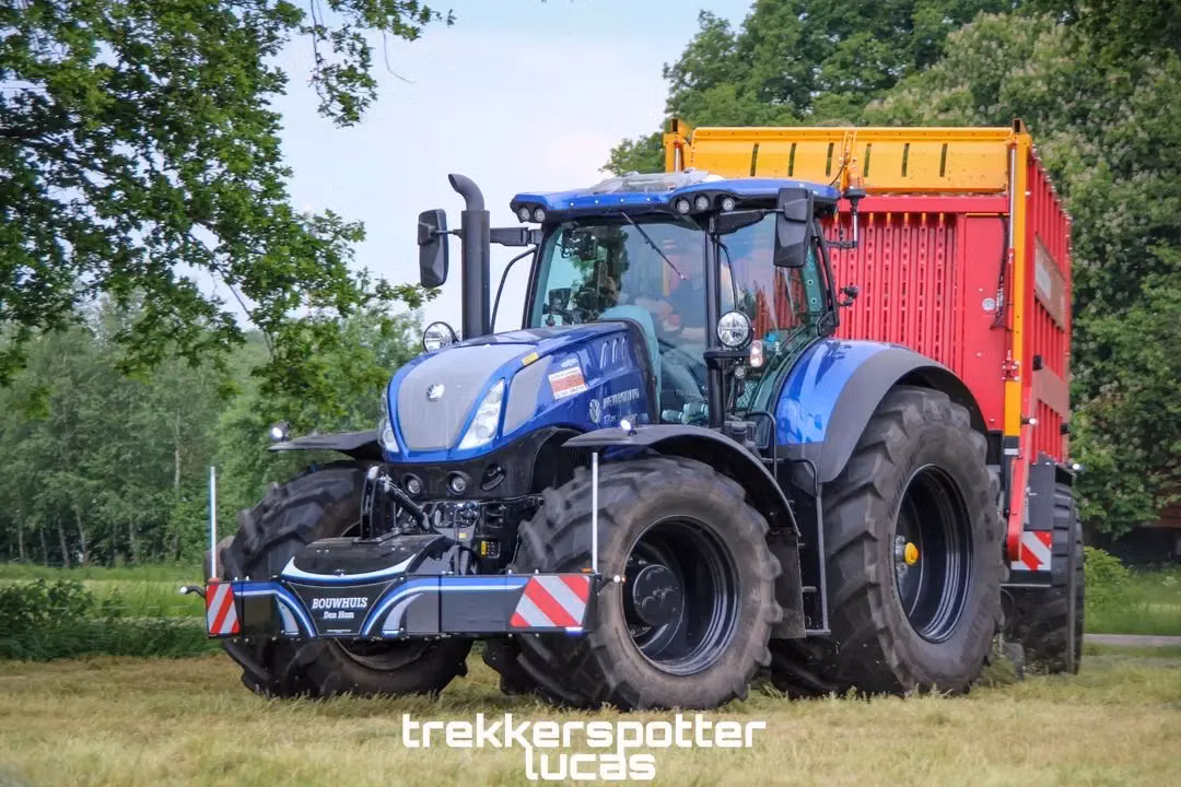 Blue tractor with front weight bumper and large rear trailer on grass, trees in background, text overlay at bottom