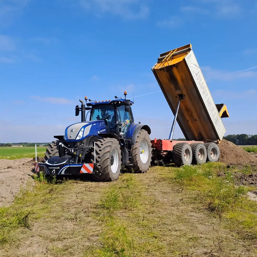 A blue tractor with a front weight bumper is pulling a large yellow tipper trailer unloading soil on a field