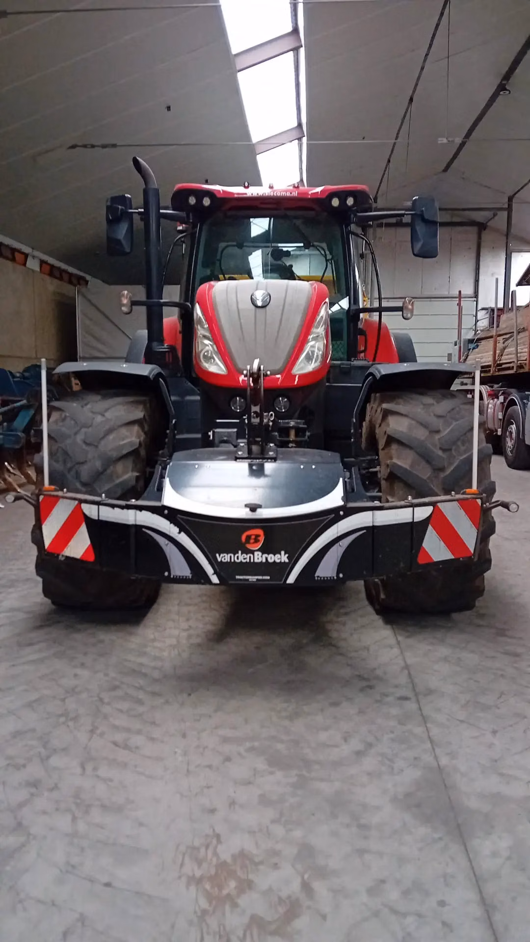 A red tractor with large tires fitted with a vandenBroek front weight bumper and reflective safety markers inside a building