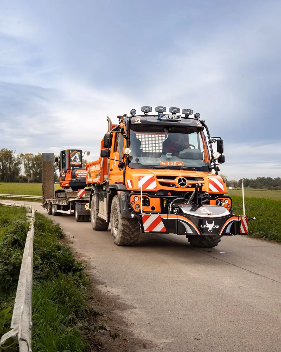 Orange utility vehicle with large front weight bumper and reflective stripes, towing a trailer on a rural road