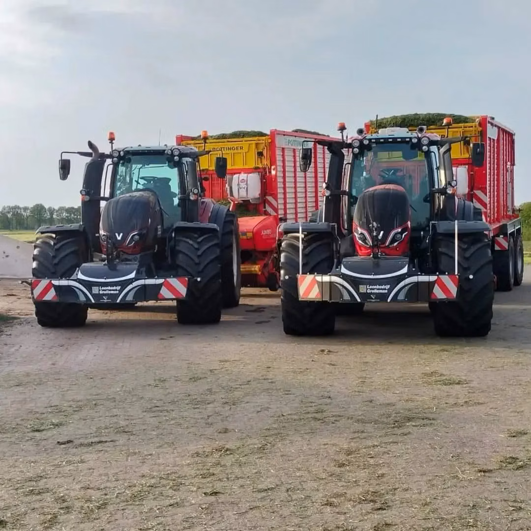 Two black tractors with front weight bumpers are parked side by side, each attached to a large red and yellow trailer