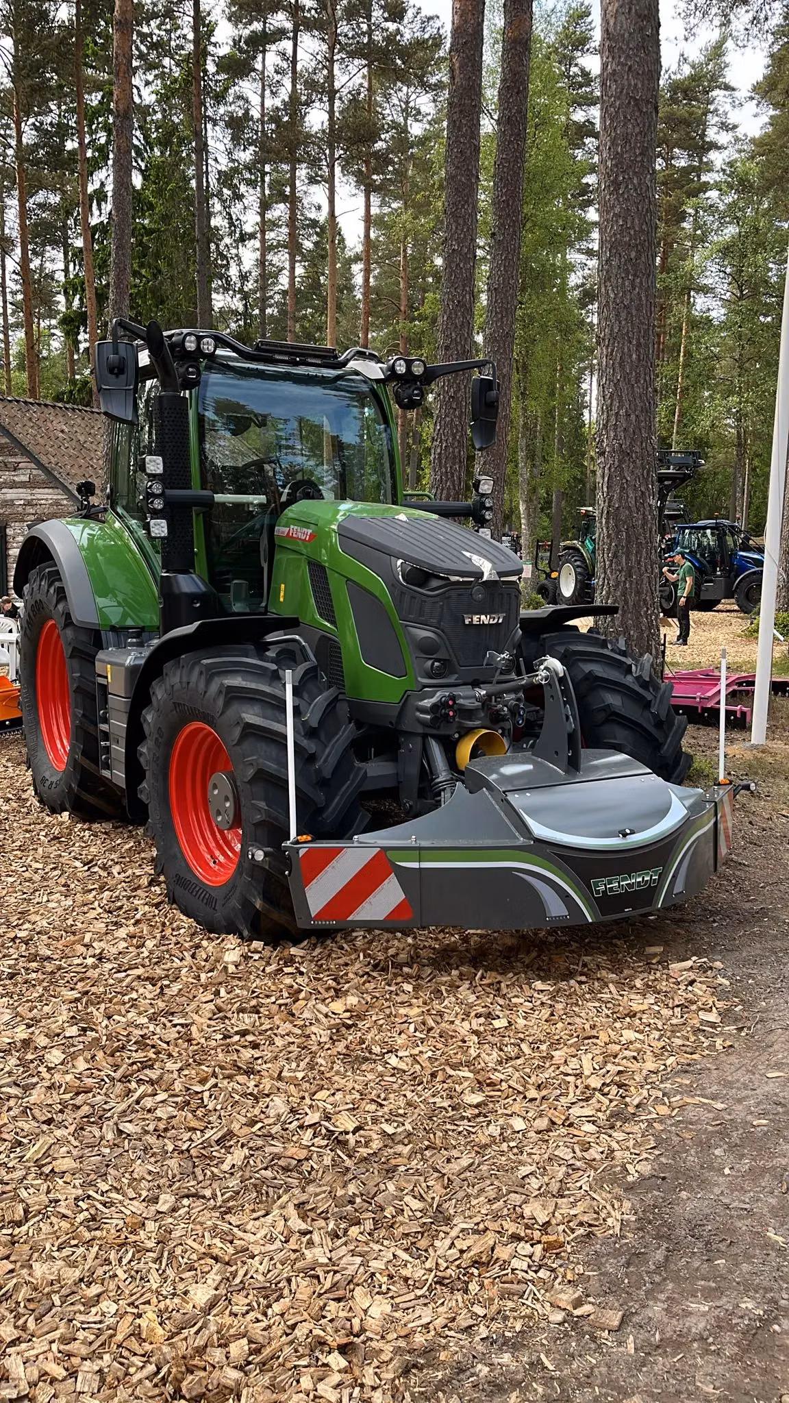 Green tractor with a front weight bumper attachment featuring red and white reflective stripes, parked on wood chips