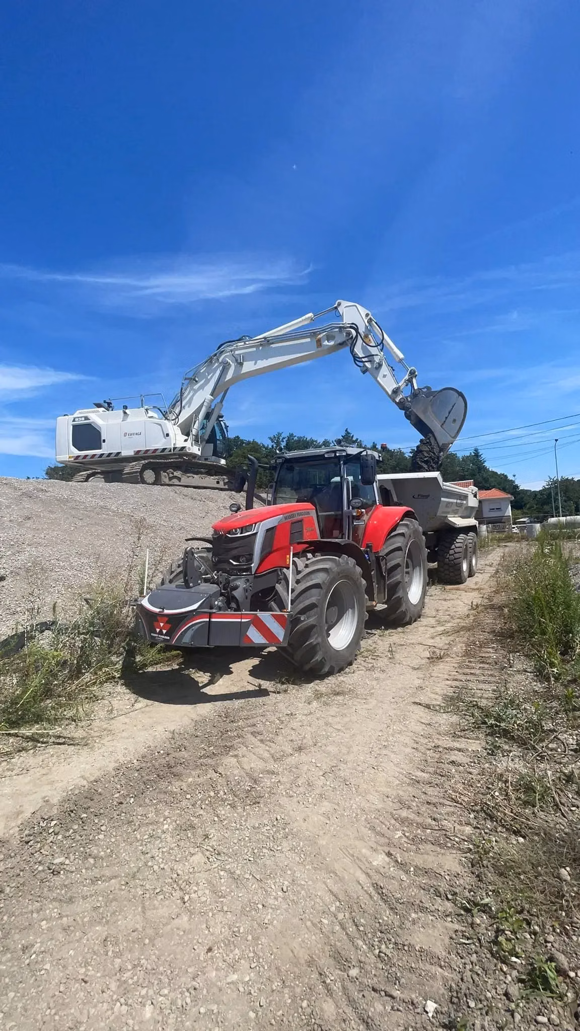 A red tractor with a front weight bumper is parked on a dirt road near construction equipment and a dump truck