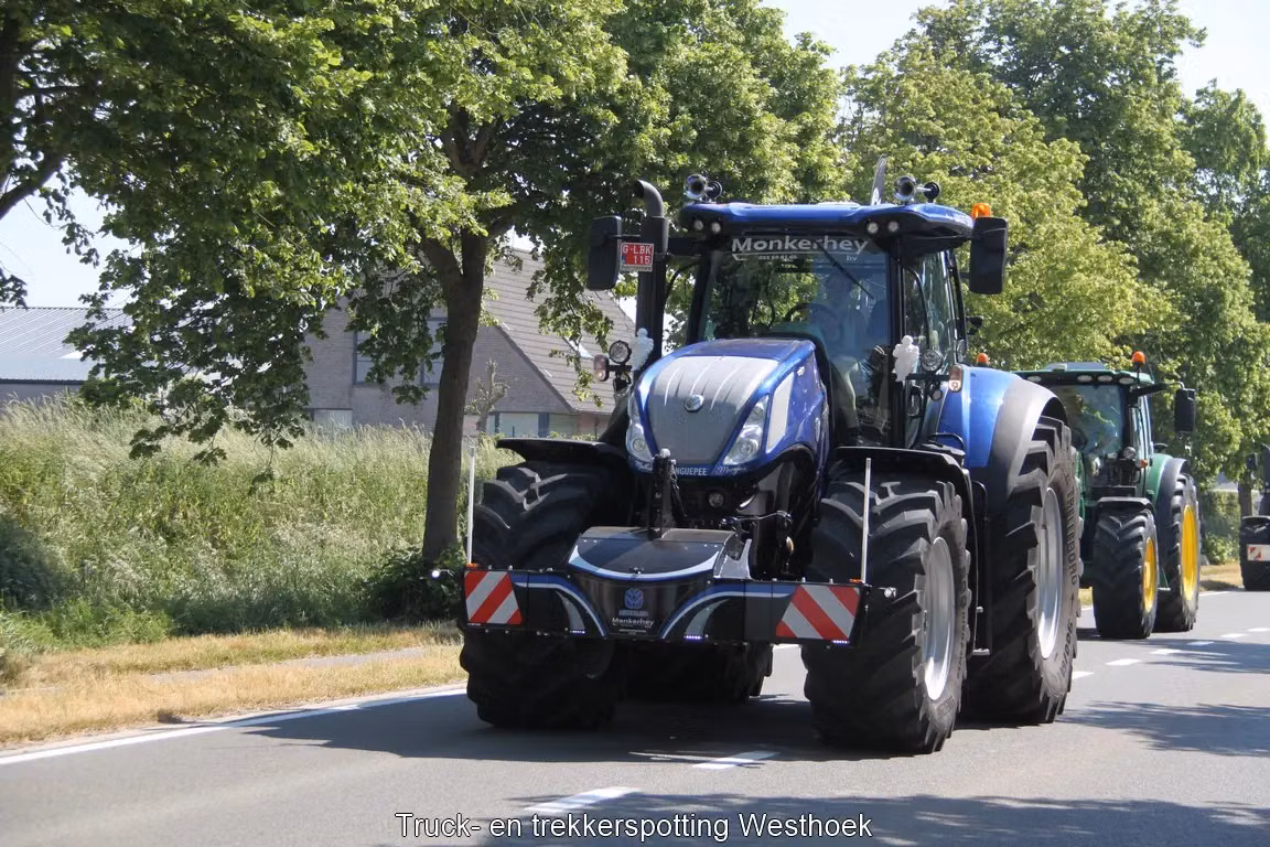 A blue tractor drives on a road with a front weight bumper attached, followed by another tractor in the background