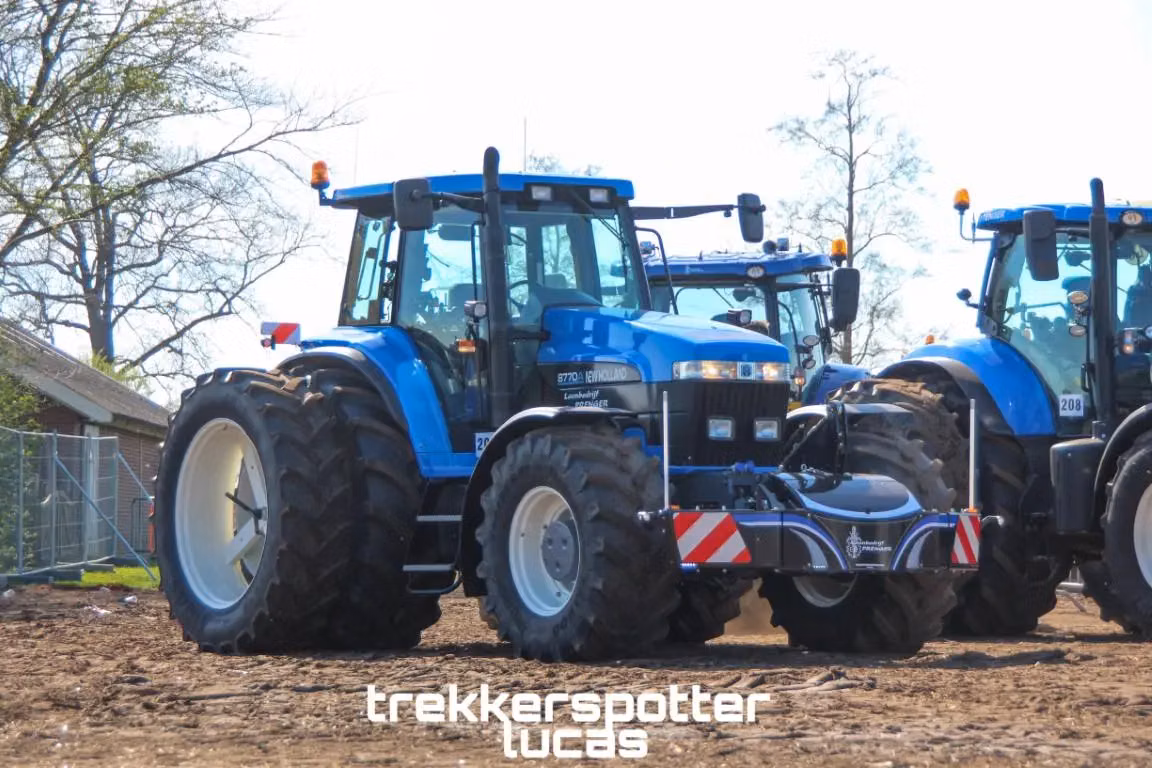 Blue tractor with a front weight bumper featuring red and white safety stripes on a dirt lot with other tractors nearby