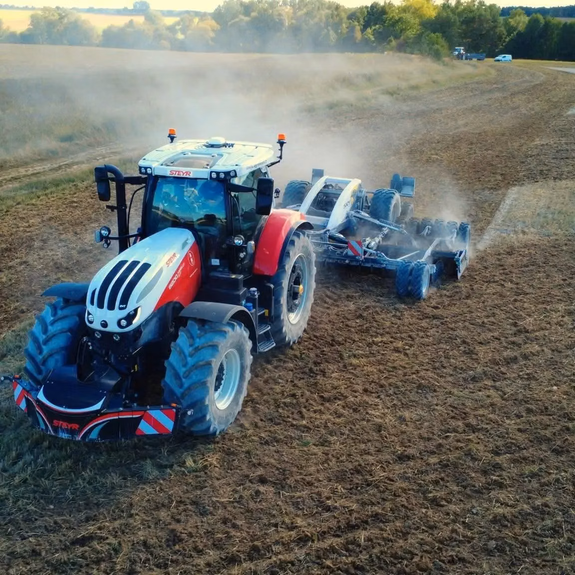 A tractor with a front weight bumper and LED lights works in a dusty field, pulling a large cultivator attachment