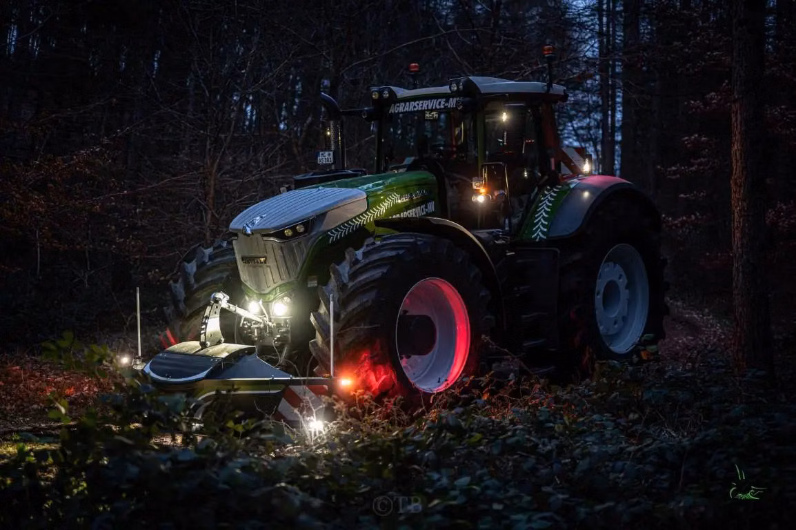 Green tractor with illuminated front weight bumper attachment on a forest path at dusk, lights glowing warmly