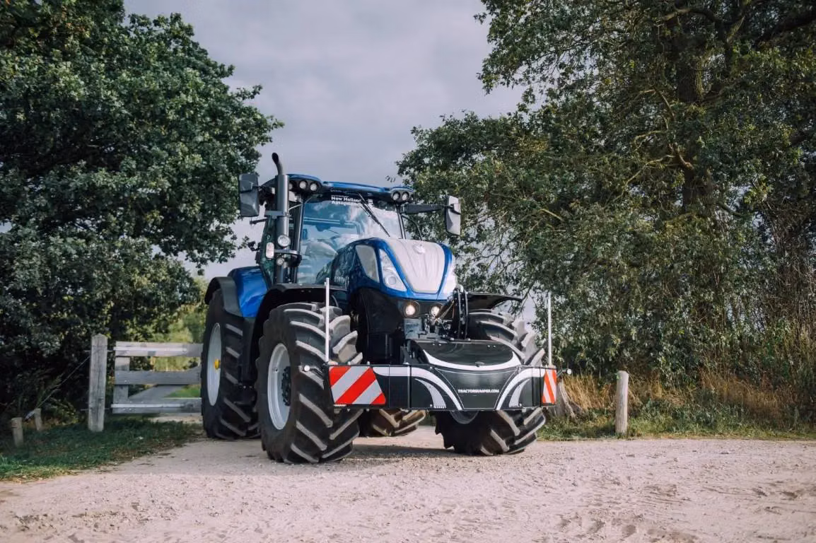 A blue tractor fitted with a black front weight bumper featuring red and white reflective safety markings.