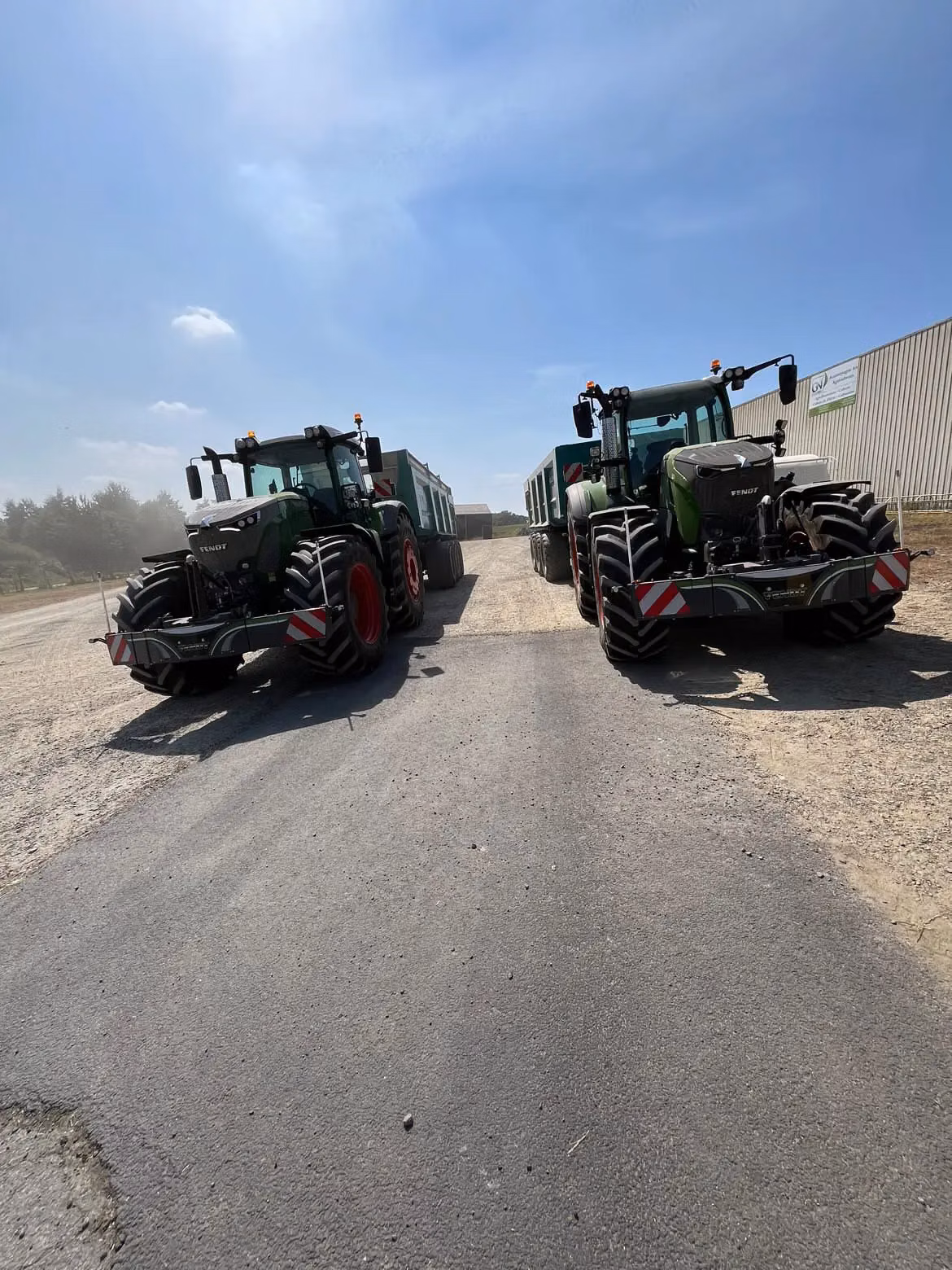 Two green tractors with front weight bumpers and large trailers are parked on a gravel and asphalt surface under a blue sky