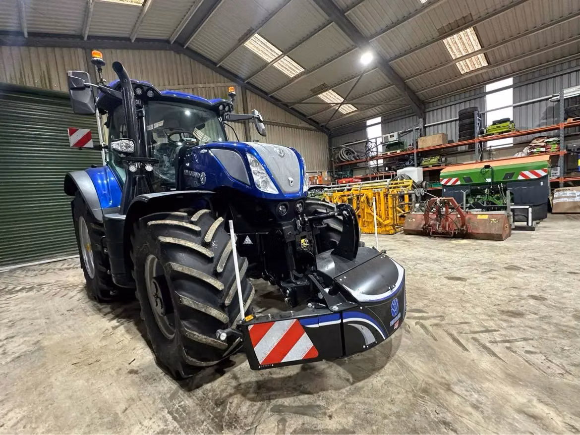 A blue tractor in a shed fitted with a front weight box featuring LED lights and red-white hazard stripes