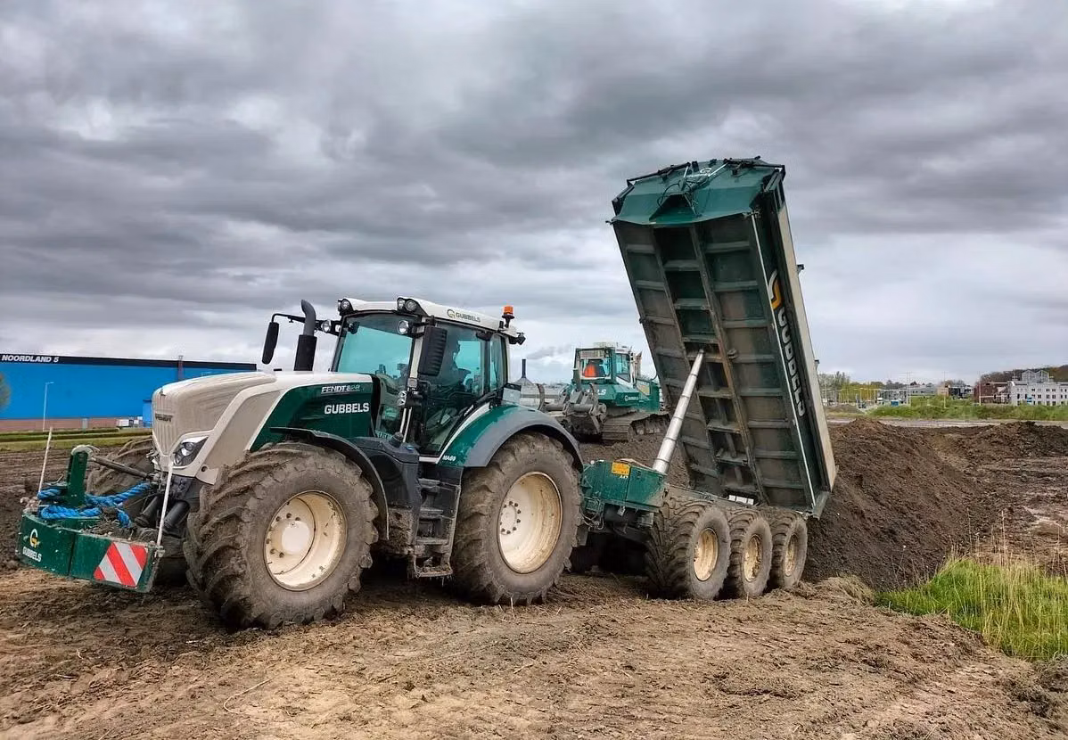 Tractor with a front weight bumper and LED lighting attached, hitched to a raised dump trailer on a muddy construction site