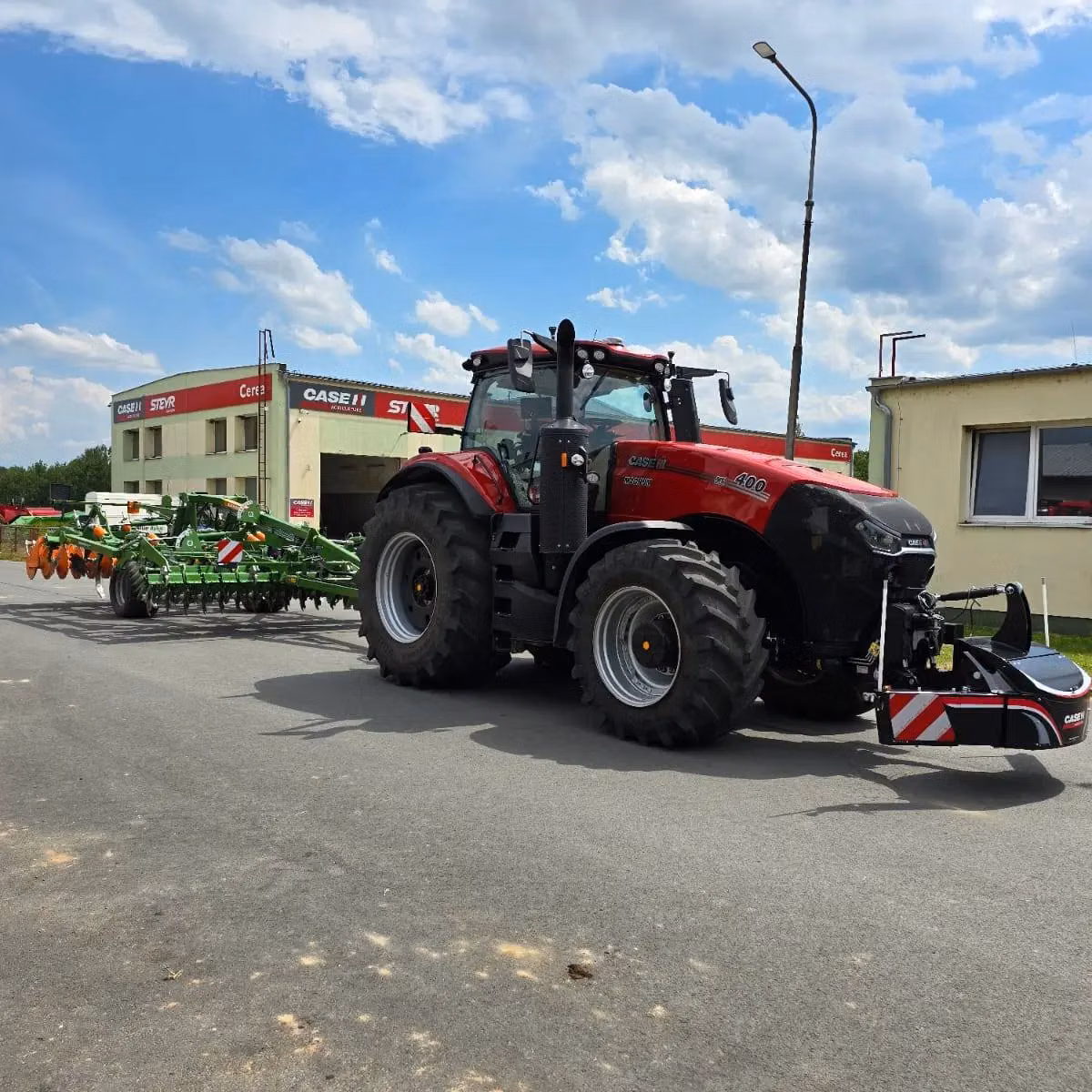 A red tractor with large tires and a front weight bumper is parked on pavement with a green implement attached at the rear