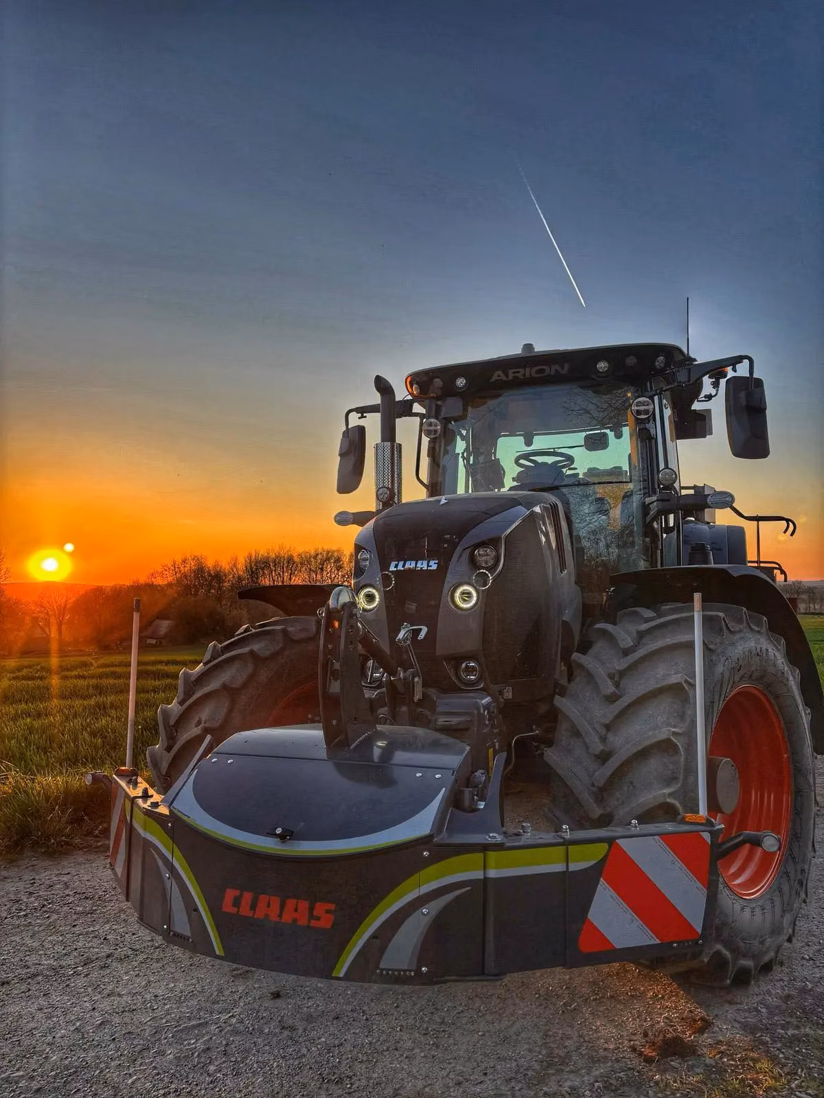 A tractor with a CLAAS front weight bumper is parked on a dirt path at sunset with green fields in the background