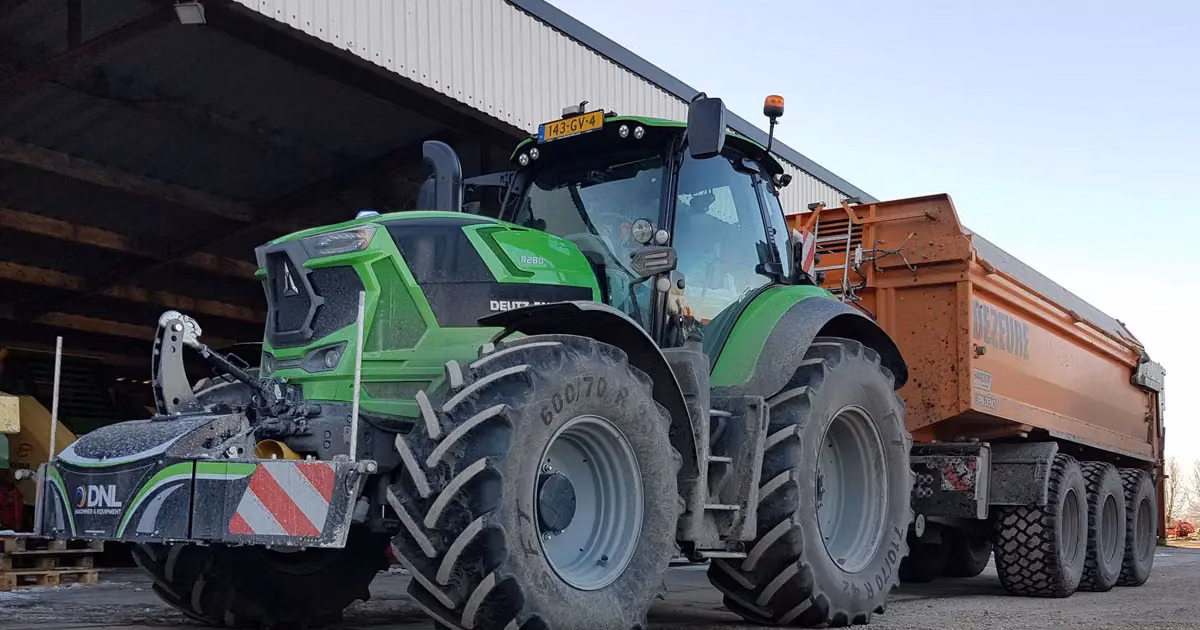 Green tractor with a front weight bumper and large trailer parked near a shed on a farmyard