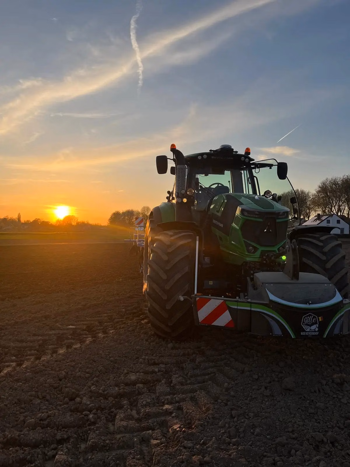 A tractor with a front weight bumper is parked on a field at sunset with visible tire tracks on the soil