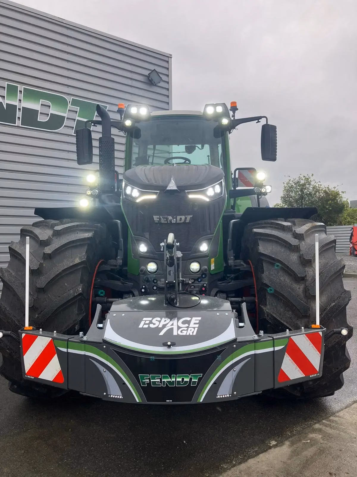 Large tractor with a front weight bumper featuring Espace Agri and Fendt branding, parked outside a building