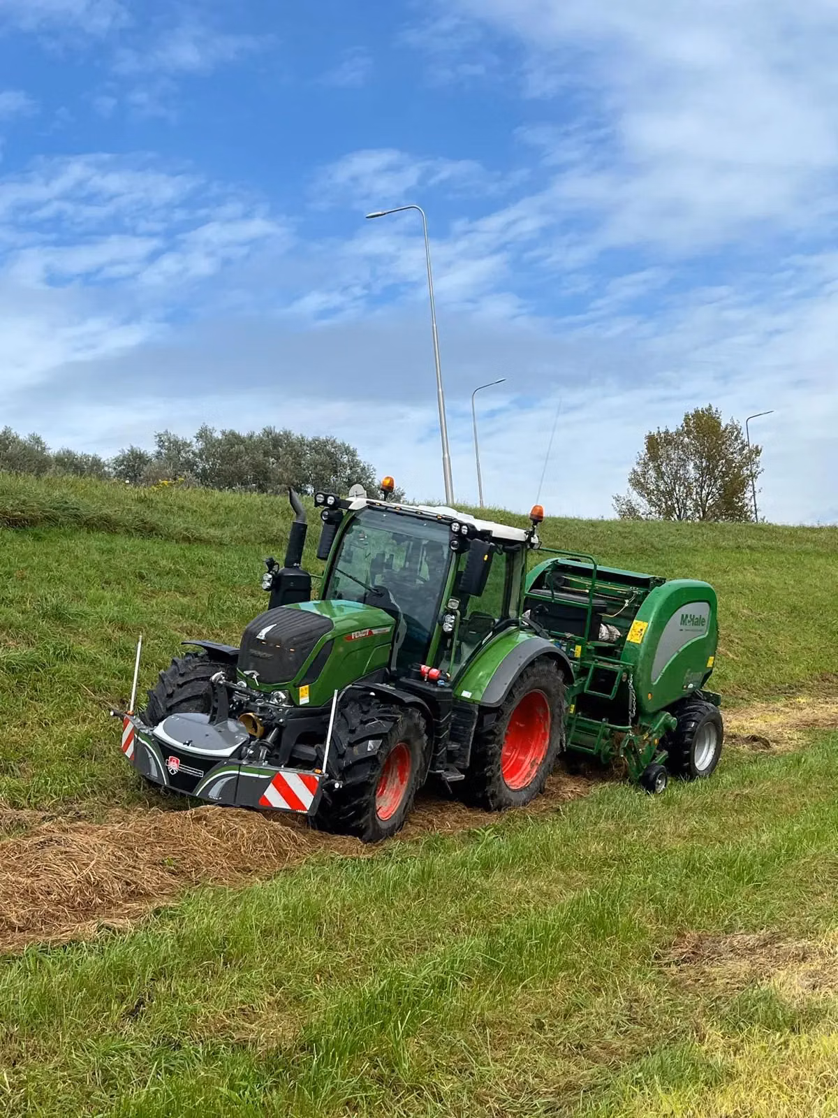 Green tractor on grass slope with a weighted front bumper and attached green round baler, under a partly cloudy sky