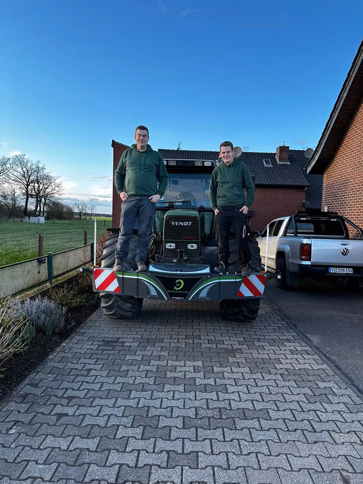 Two people in green hoodies stand on a tractor equipped with a front weight bumper in a paved driveway