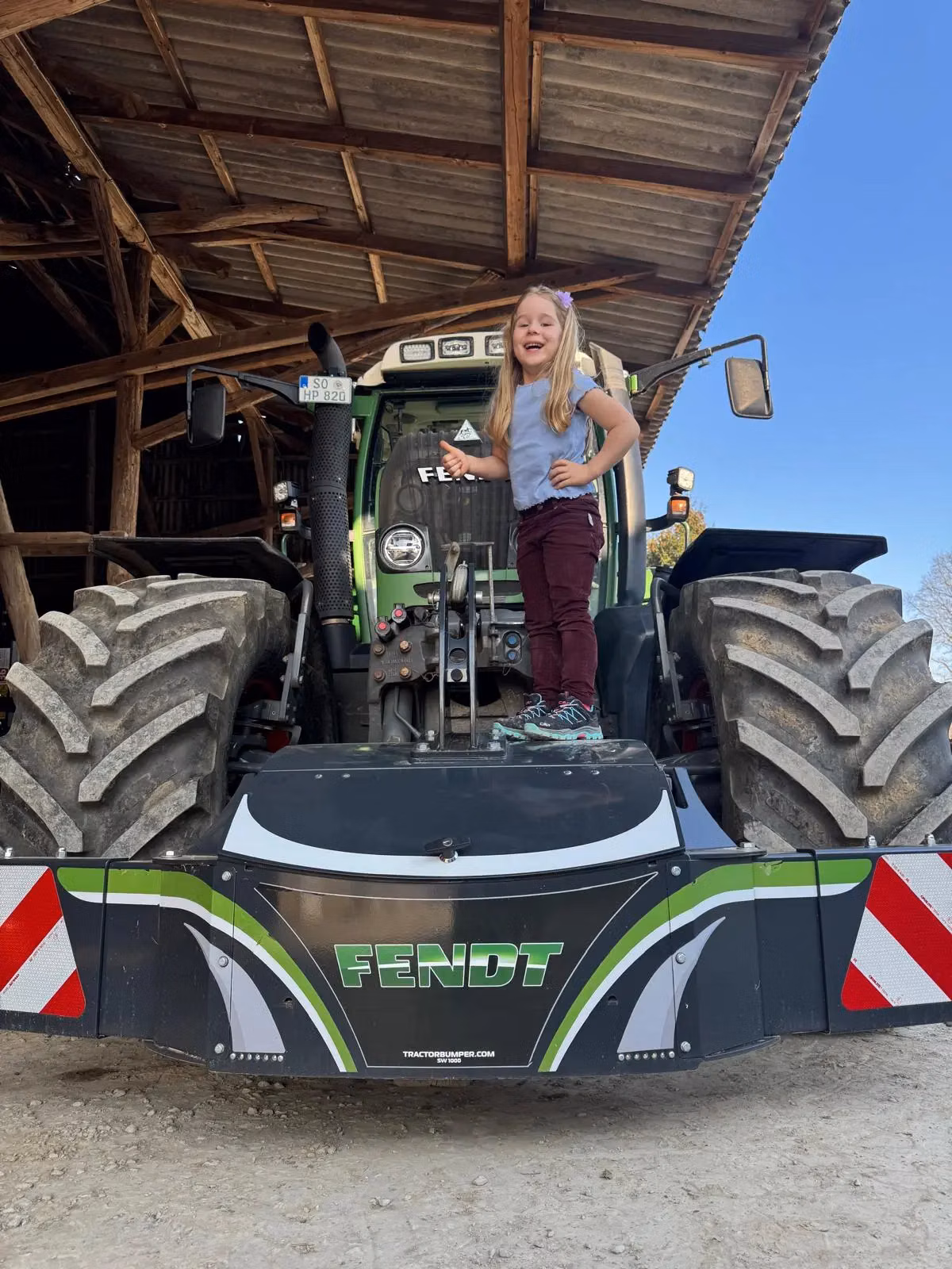 Smiling child stands on a Fendt front weight bumper attached to a tractor in an open-sided shed.