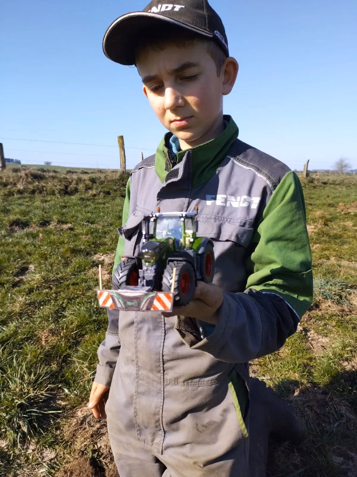 A boy outdoors in work clothes holds a toy tractor with a front weight bumper in his hand