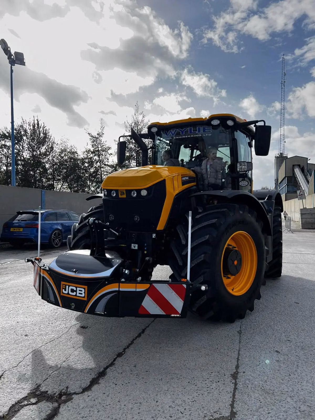 Yellow and black tractor with a front weight bumper labeled JCB, parked on concrete with two people in the cab
