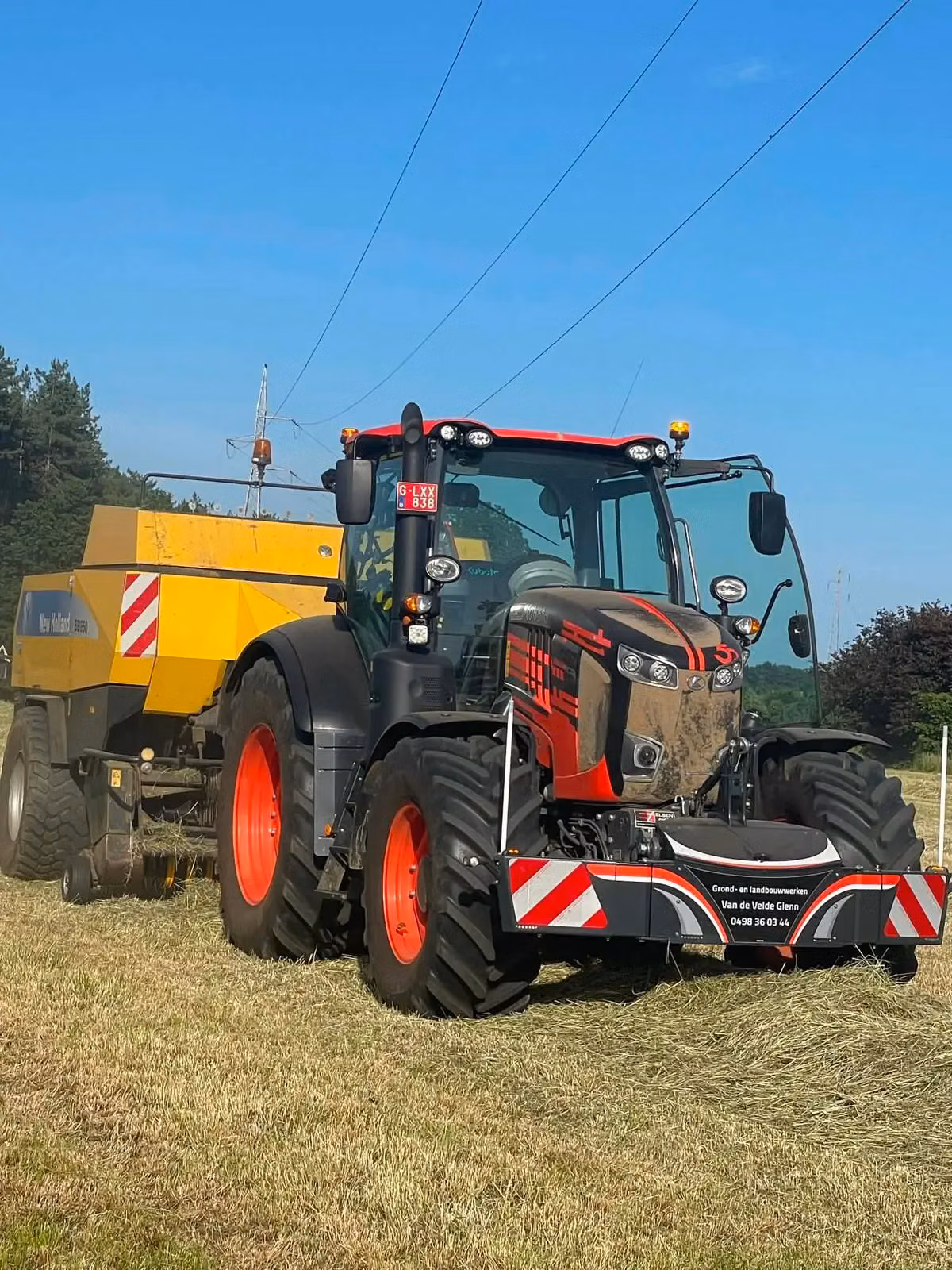 A tractor with a front weight bumper and red reflective stripes is working in a field, attached to a yellow baler.