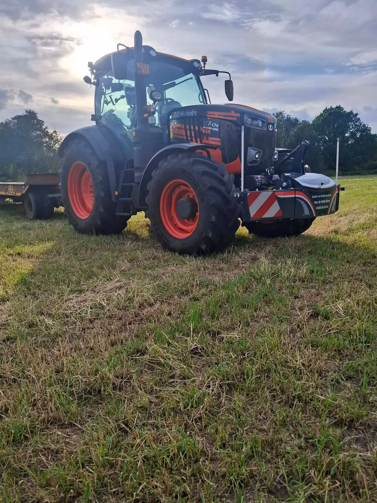 A tractor with a front weight bumper is parked on grass with a trailer attached in the background