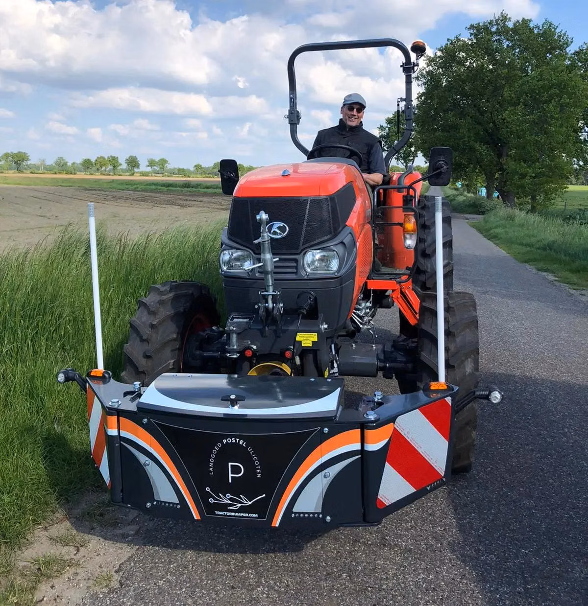 A person driving an orange tractor fitted with a black and orange front weight bumper on a rural road
