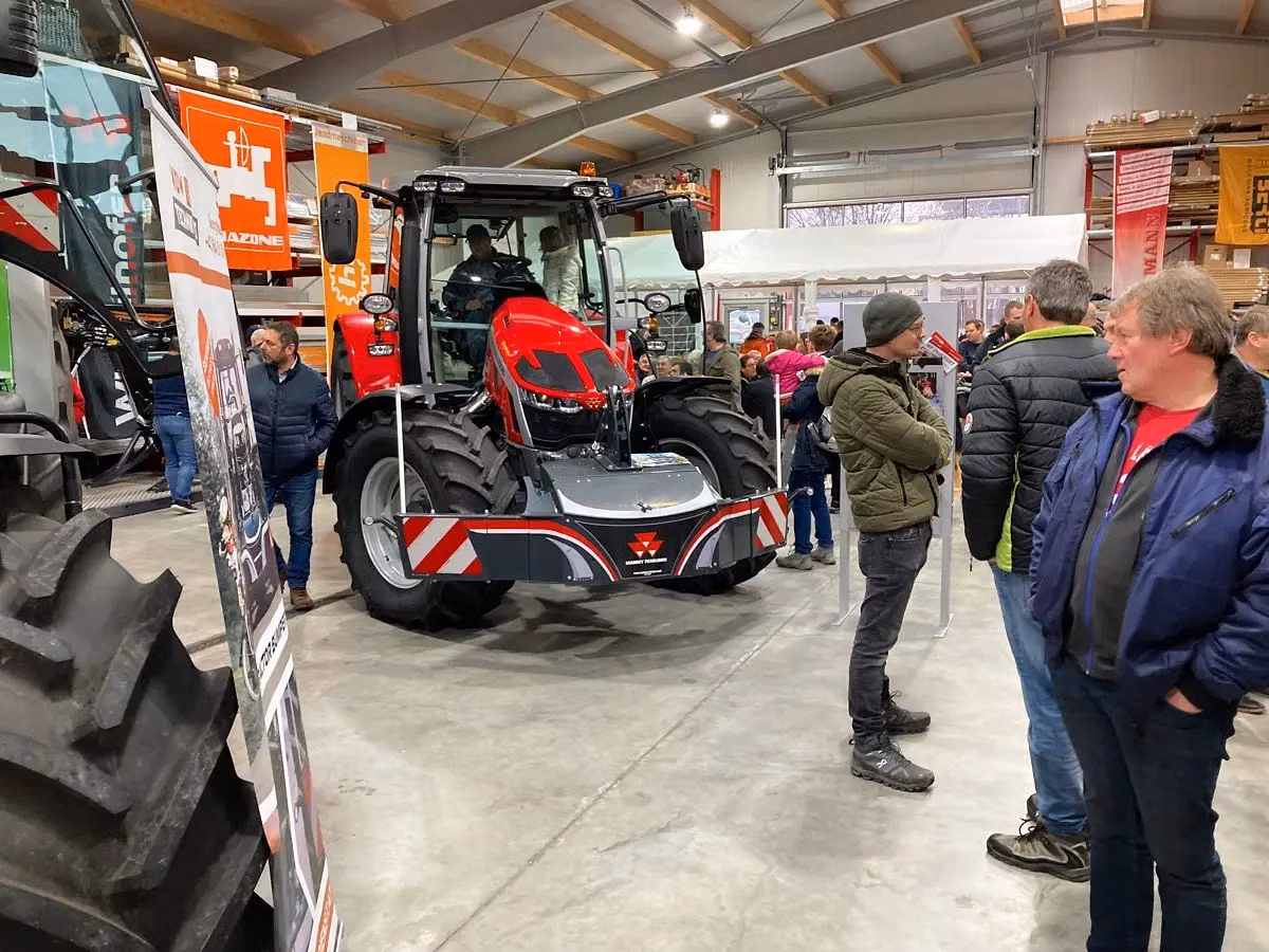 A red tractor with a front weight bumper is displayed indoors with people standing and walking around.
