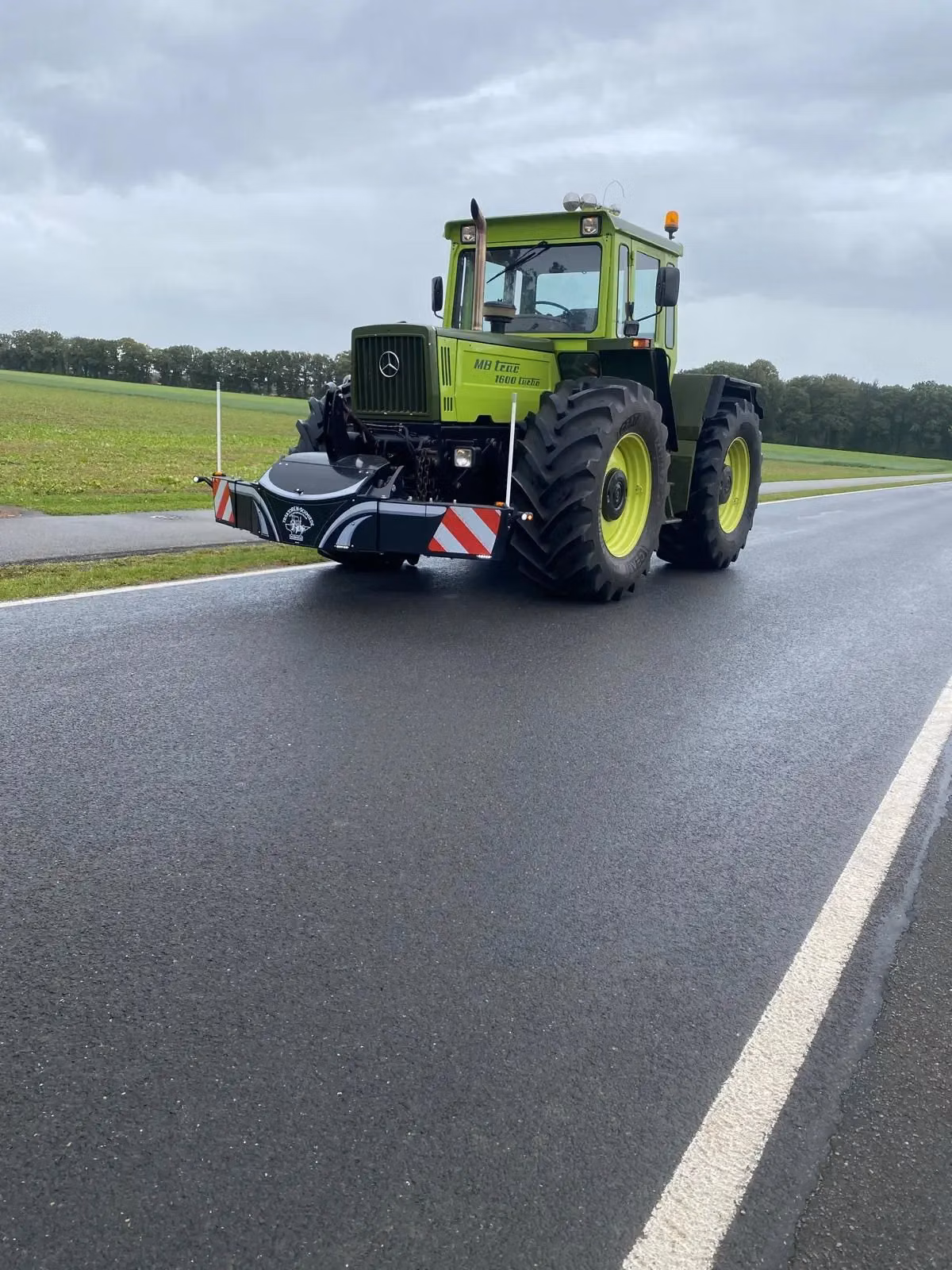 Green tractor with a large front weight bumper featuring red and white warning stripes on a wet road