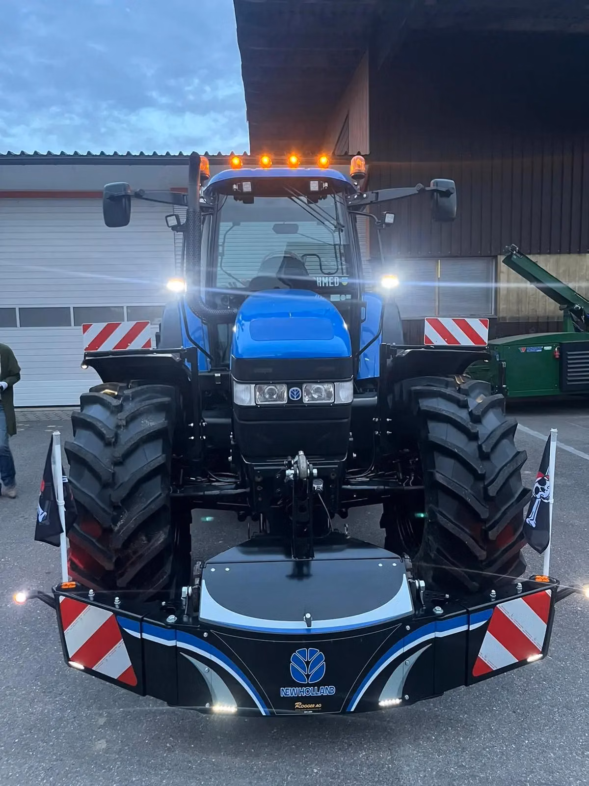 Blue tractor with large front wheels, orange lights on top, and a New Holland branded front weight bumper attached