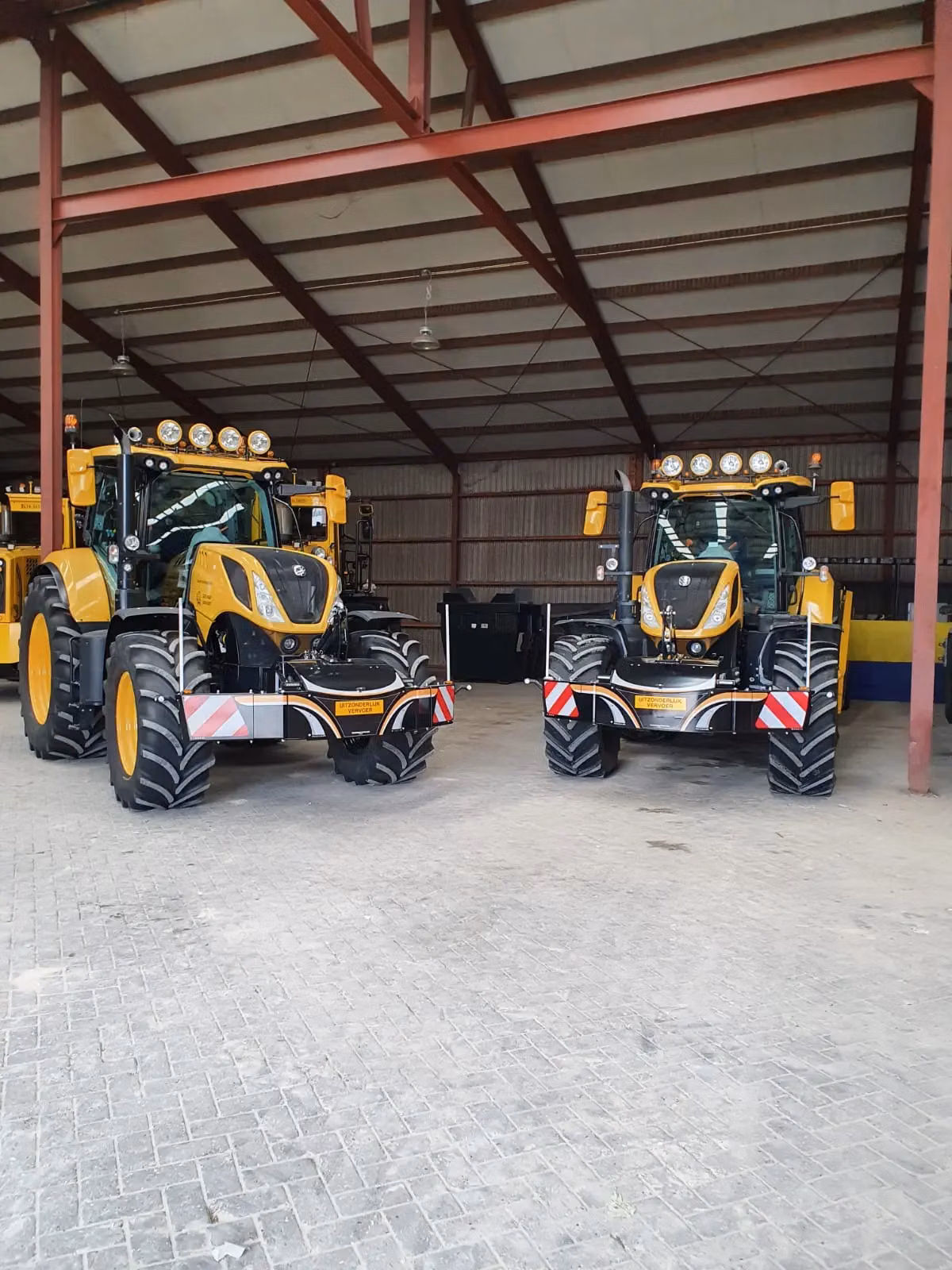 Two yellow tractors indoors each fitted with a front weight bumper with red and white safety markings